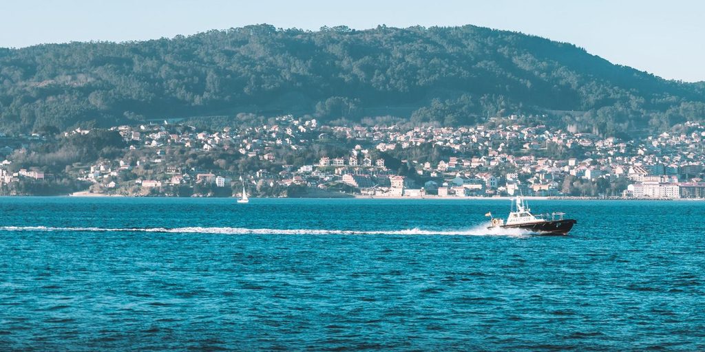 Barco navegando por una ría de las Rías Baixas con casas y monte al fondo